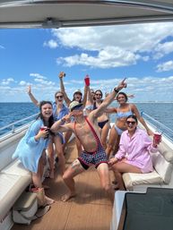Energetic group of friends partying on a sunlit yacht in open coastal waters, wearing swimsuits and holding red cups under a blue sky with scattered clouds.