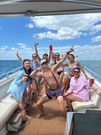 Energetic group of friends partying on a sunlit yacht in open coastal waters, wearing swimsuits and holding red cups under a blue sky with scattered clouds.