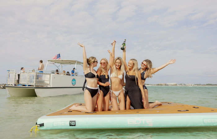 Five friends in swimsuits cheering on a floating platform in shallow turquoise coastal waters beside a party boat, one holding a champagne bottle.