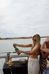 Smiling friends pop champagne on a coastal boat deck near a sandy shoreline, orange juice and a bowl on the table for mimosas.