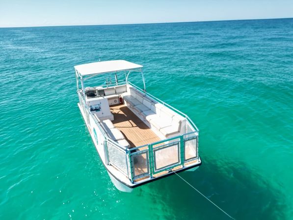 Aerial view of a white pontoon boat with cushioned seating anchored in crystal-clear turquoise ocean on a sunny day