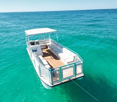 Aerial view of a white pontoon boat with cushioned seating anchored in crystal-clear turquoise ocean on a sunny day