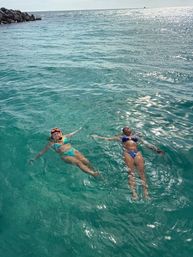 Two swimmers in bikinis — one with snorkel gear — floating on their backs in clear turquoise ocean near a rocky jetty on a sunny tropical day.