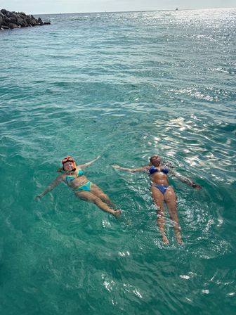 Two swimmers in bikinis — one with snorkel gear — floating on their backs in clear turquoise ocean near a rocky jetty on a sunny tropical day.