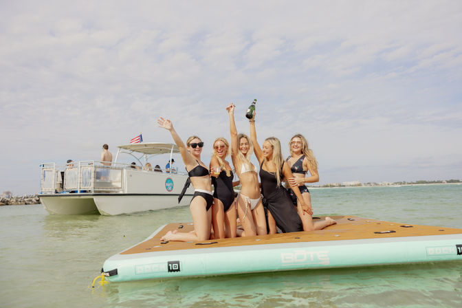 Five women in swimsuits cheering and holding a champagne bottle on a large inflatable dock near a party boat in shallow turquoise coastal water under a sunny sky