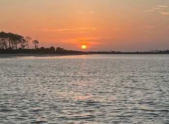 Golden sunset over a coastal bay, the sun sinking behind a low tree-lined shoreline with orange sky reflected on rippling water.