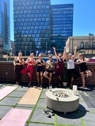Rooftop yoga class with seven people in colorful activewear holding tree pose on mats on a sunny urban rooftop, modern glass office buildings forming a downtown skyline behind them.