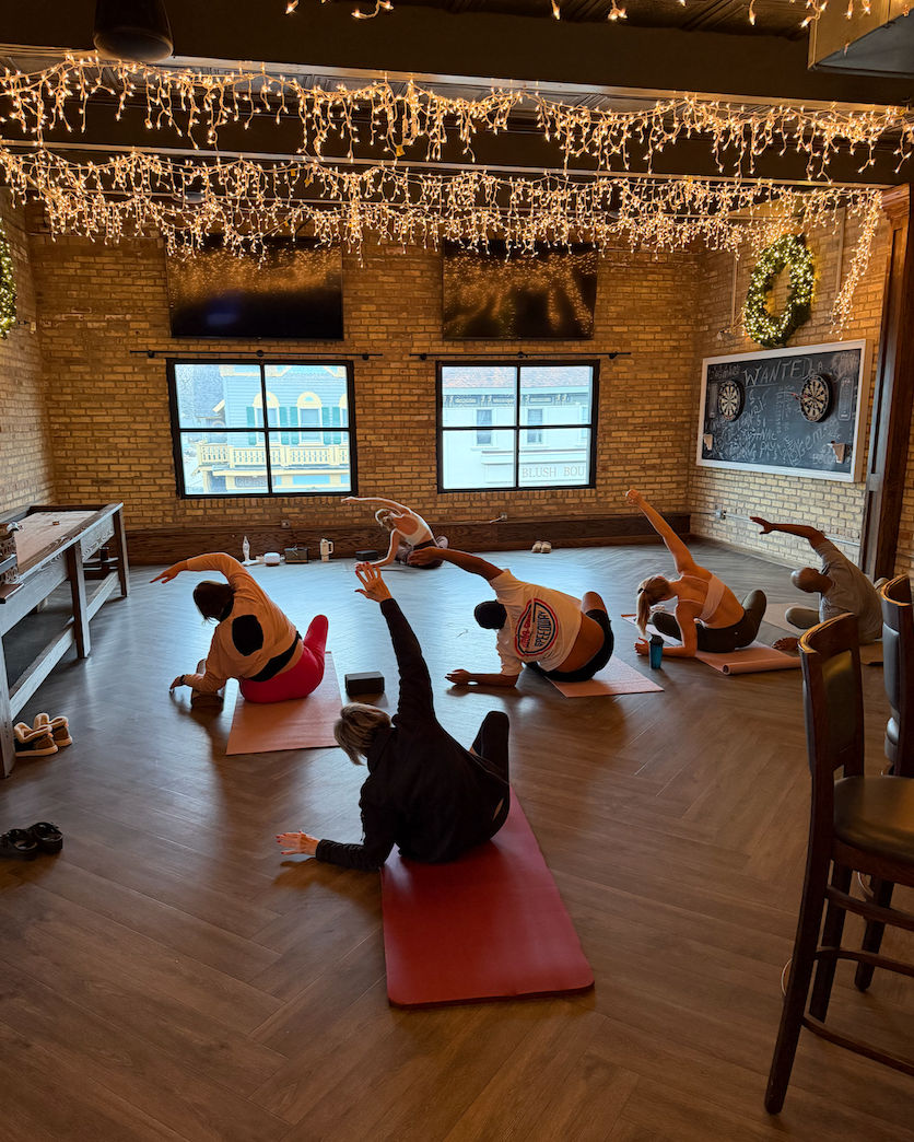Group yoga/stretch class in a cozy loft-style studio with exposed brick walls, herringbone wood floor, twinkling string lights, large windows overlooking a downtown street, chalkboard wall and participants on mats doing side stretches.