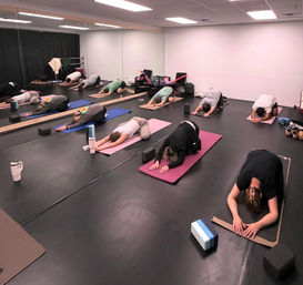 Group yoga class in an indoor studio: students on mats in child's pose with blocks, water bottles, and a mirrored wall