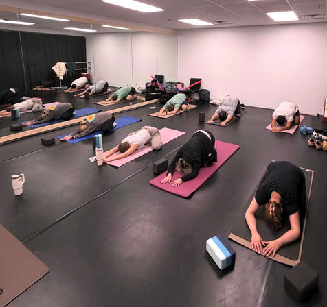Group yoga class in an indoor studio: students on mats in child's pose with blocks, water bottles, and a mirrored wall