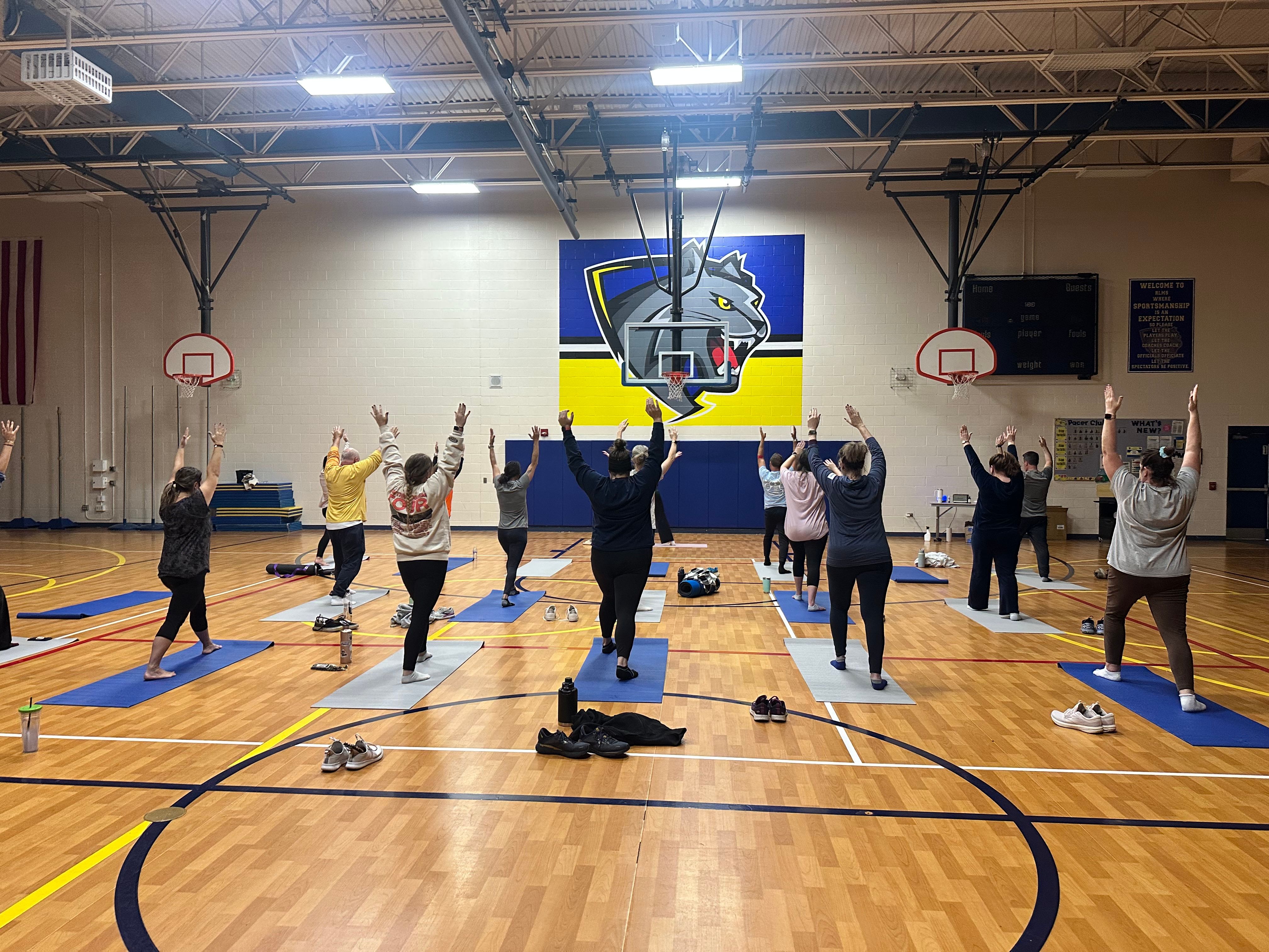 Group yoga class on mats in a school gymnasium, participants raising arms toward a large wolf mascot mural above basketball hoops on the hardwood court.