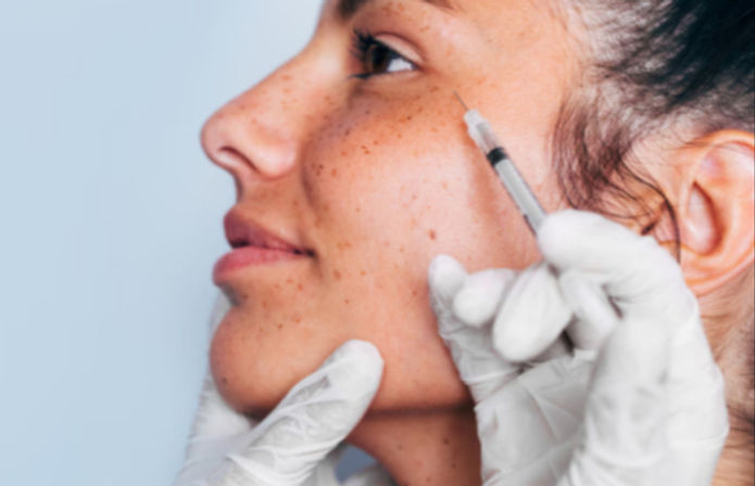 Close-up profile of a freckled face receiving a cosmetic cheek/under-eye injection from a gloved clinician in an aesthetic clinic.