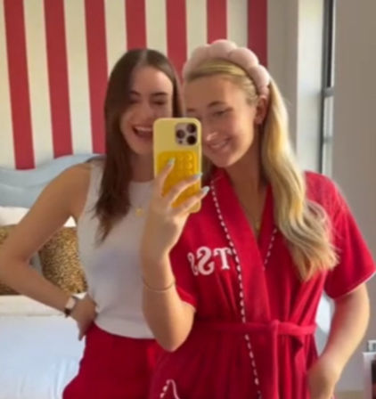 Two smiling friends taking a mirror selfie in a bedroom with red-and-white striped wall; one wearing a red robe and padded headband, the other in a white tank top holding a yellow phone case.