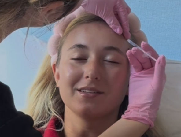 Relaxed woman with fluffy headband, eyes closed and smiling as a practitioner in pink gloves administers a cosmetic forehead injection with a syringe during an aesthetic clinic treatment.