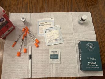 Top-down view of an at-home skincare/medical treatment setup on a wooden table: orange-capped syringes, vials, sterile gauze and non-woven sponge packets, a peel prep towelette, a post-peel aftercare kit box, and a sharps container arranged on a white disposable medical pad.
