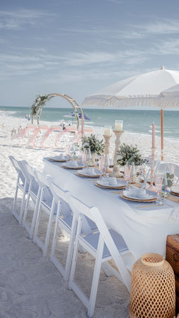 Oceanfront beach wedding reception set on white sand with folding chairs, linen-covered table, floral centerpieces, candles, parasol and wooden ceremony arch by turquoise sea under a clear sky