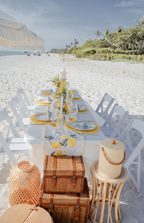 Lemon-themed beach table setting on white-sand shore—long table with white folding chairs, yellow citrus runner, woven placemats, glassware and candles under a fringed umbrella, wicker trunks and palm-lined coastline in the background.