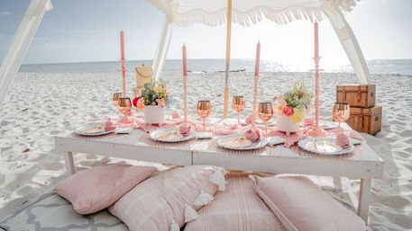 Sunlit boho-chic beach picnic on white sand: low rustic table under a fringe canopy with pink taper candles, rose-gold goblets, floral centerpieces, wicker baskets and plush cushions overlooking the ocean.