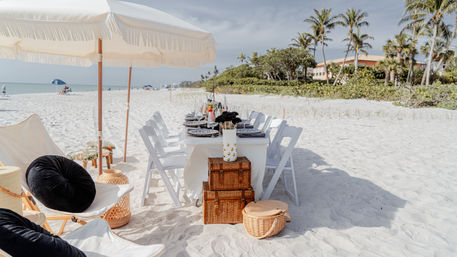 Elegant beach dining setup on white sand with a fringe umbrella, long white-table and chairs, wicker baskets, palm trees and ocean view