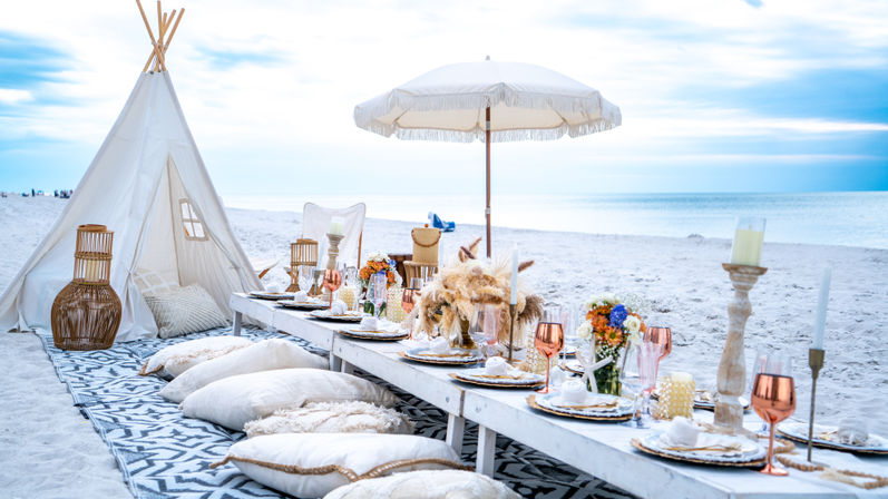 Boho-chic beach picnic on white sand with a low rustic table set with plates, copper goblets, floral centerpieces and candles, surrounded by floor cushions, a fringe umbrella and canvas teepee with the ocean horizon in the background.