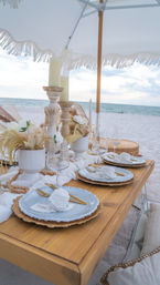 Seaside beach dinner setup on white sand under a fringed umbrella — low wooden table with layered plates, gold cutlery, wine glasses, candles and neutral dried floral accents facing the ocean