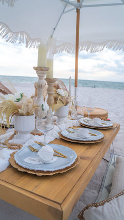 Seaside beach dinner setup on white sand under a fringed umbrella — low wooden table with layered plates, gold cutlery, wine glasses, candles and neutral dried floral accents facing the ocean
