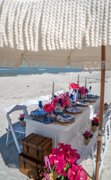 Oceanfront beach tablescape under a fringed umbrella — white folding chairs, blue-and-white plates, pink floral centerpieces and wicker trunks on sandy shore