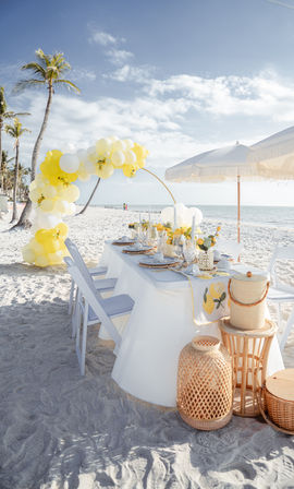 Sunlit tropical beach scene with a white-linen dining table set for a seaside celebration, lemon-themed place settings, rattan accents, yellow-and-white balloon arch, palm trees and calm ocean under a blue sky.