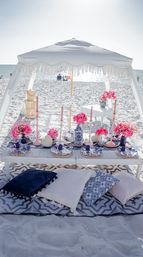 Oceanfront beach picnic under a white fringe canopy: low wooden table set with blue-and-white plates, woven chargers, pink flowers and taper candles, and assorted throw pillows on a patterned rug on the sunlit sandy shore.