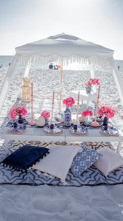 Oceanfront beach picnic under a white fringe canopy: low wooden table set with blue-and-white plates, woven chargers, pink flowers and taper candles, and assorted throw pillows on a patterned rug on the sunlit sandy shore.