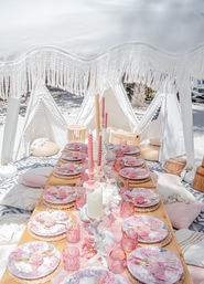 Beachside boho picnic setup on sand with a low wooden table under a white fringed canopy, pastel pink floral plates on woven chargers, pink glassware and twisted taper candles, surrounded by cushions and small white teepee tents