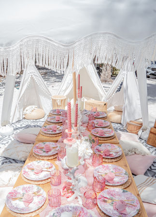 Beachside boho picnic setup on sand with a low wooden table under a white fringed canopy, pastel pink floral plates on woven chargers, pink glassware and twisted taper candles, surrounded by cushions and small white teepee tents