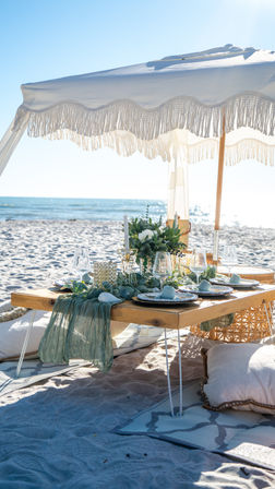 Sunlit boho beach picnic on sandy shore with a low wooden table under a white fringed umbrella, floral centerpiece, candles, glassware and cushions overlooking the sparkling ocean