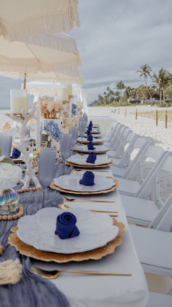 Coastal-chic beachside table on white sand with fringed umbrellas, white plates on gold chargers, navy napkin roses, textured blue glassware, candle and coral decor, and palm trees along the tropical shoreline.