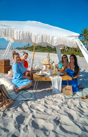 Group of four friends enjoying a stylish beach picnic under a white fringed canopy on a sunny sandy beach with a low wooden table, place settings, and palm trees