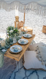 Boho beach picnic on a sandy coast: low wooden table under a fringed umbrella with a floral centerpiece, blue-gray place settings, gold cutlery, wine glasses, woven lanterns and tassel cushions.