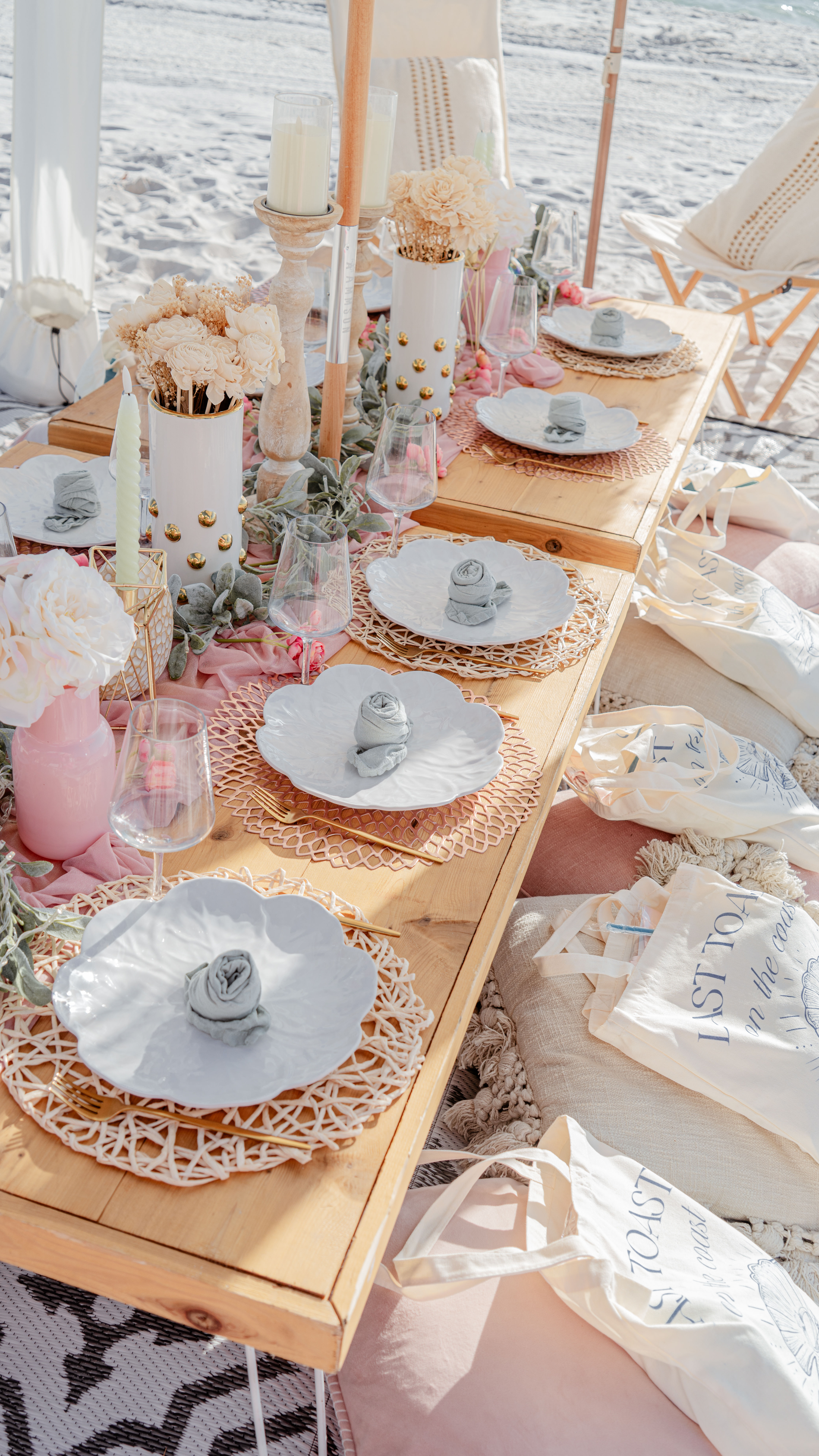 Sunlit boho seaside picnic on white sand — low wooden table set with scalloped white plates on woven chargers, rolled napkin roses, gold cutlery, pink-and-cream floral arrangements, candles, glassware and cushioned beach seating with tote bags.