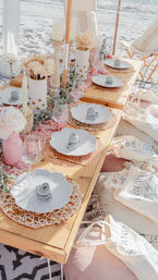 Sunlit boho seaside picnic on white sand — low wooden table set with scalloped white plates on woven chargers, rolled napkin roses, gold cutlery, pink-and-cream floral arrangements, candles, glassware and cushioned beach seating with tote bags.