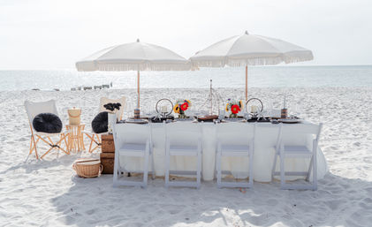 Sunlit oceanfront beach dining setup on white sand: long white-clothed table with six folding chairs, two fringed umbrellas, a small wooden sailboat centerpiece and colorful floral arrangements, calm sea on the horizon.