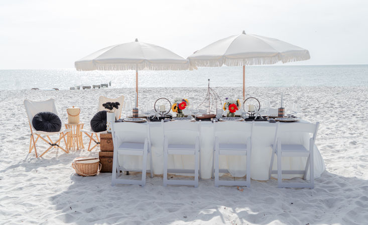Sunlit oceanfront beach dining setup on white sand: long white-clothed table with six folding chairs, two fringed umbrellas, a small wooden sailboat centerpiece and colorful floral arrangements, calm sea on the horizon.