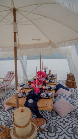 Boho beach picnic under a fringed white canopy on a sandy shore, featuring a low wooden table set with blue-and-white plates, navy runner, pink floral centerpieces, rose-gold goblets, tall candles, floor cushions and woven baskets, with the ocean in the background.