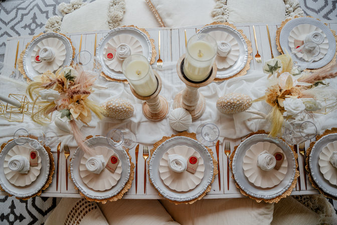Overhead view of an elegant boho-chic tablescape in neutral tones: scalloped white plates on gold chargers, rolled napkins with heart tags, gold flatware, glassware, wooden candlesticks with pillar candles and dried floral centerpieces.