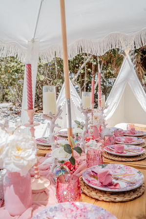 Boho pink beach table setup under a white fringed umbrella with floral plates, pink glassware, woven placemats, coral-shaped candle holders, fresh roses and white teepee tents in the background.