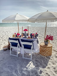 Chic beach picnic on white sand with two fringed umbrellas, a decorated table with navy cushions, pink flowers and the turquoise ocean beyond.