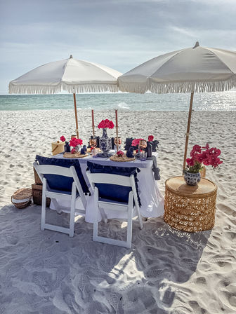 Chic beach picnic on white sand with two fringed umbrellas, a decorated table with navy cushions, pink flowers and the turquoise ocean beyond.