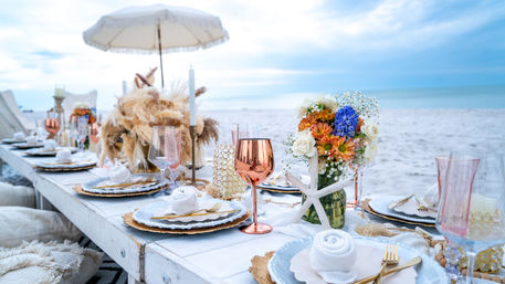 Boho-chic seaside celebration table on a white wooden beach table with gold-rimmed plates, rose-gold goblets, rolled napkins, starfish and floral centerpieces against an oceanfront horizon