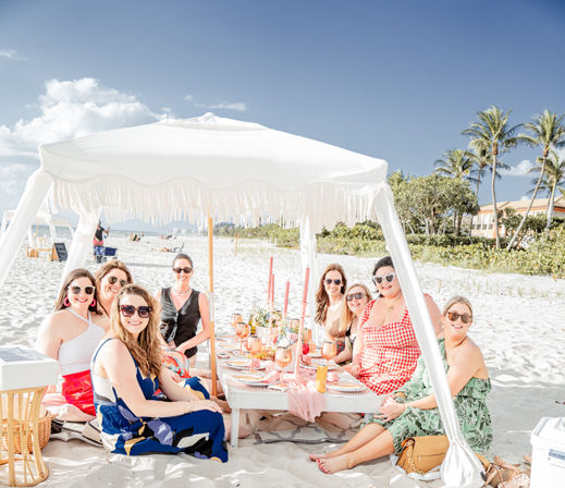 Sunny beach picnic under a white fringe canopy: group of friends seated around a low table on white sand with candles, rosé glasses and palm trees in the background.