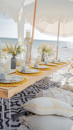 Sunlit boho seaside dining setup on a sandy beach under fringed umbrellas — low wooden table with lemon-themed plates, yellow woven placemats, glassware, floral centerpieces and cozy floor cushions overlooking the ocean.