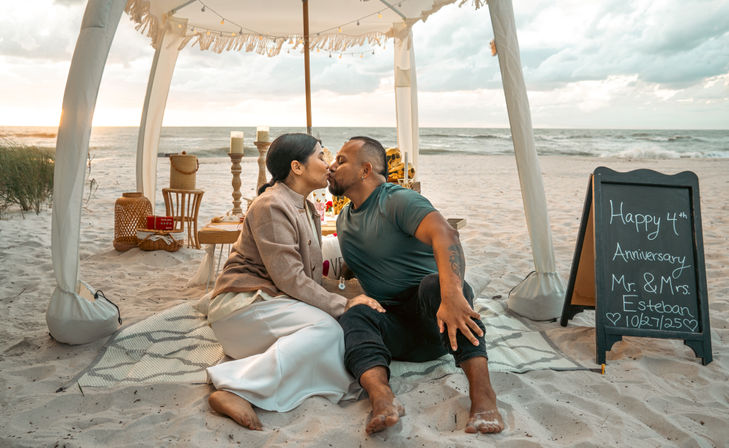 Romantic couple kissing on a sandy beach at sunset under a boho canopy with candles, picnic setup, and a chalkboard anniversary sign