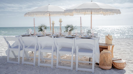 Sunlit seaside dining setup on white sand — long table with white folding chairs, fringed umbrellas, floral centerpieces, candles, glassware and wicker accents by the sparkling ocean